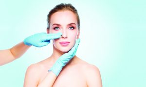 people, cosmetology, plastic surgery and beauty concept - surgeon or beautician hands touching woman face over blue background