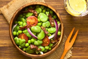 Broad bean, green pea, cherry tomato, red onion and fried bacon salad in wooden bowl, photographed overhead on dark wood with natural light