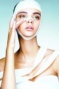 64802127 - woman removes the bandages from her head and touching her new face. portrait of young models face in medical bandage after beauty surgery. plastic surgery concept