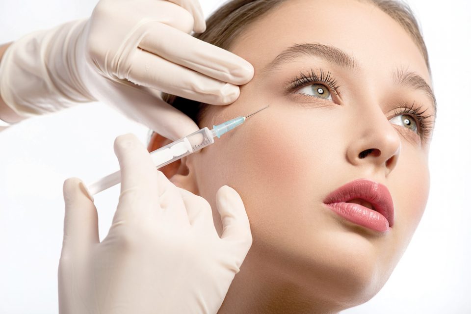 Serine young woman is getting facial botox injection. Beautician hands in gloves holding syringe near her face